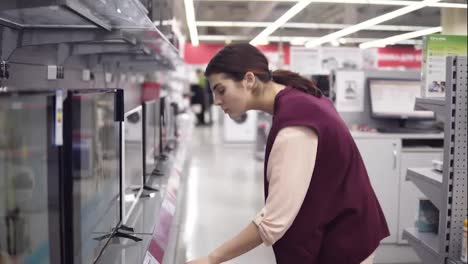 Una-Clienta-Joven-Que-Elige-Un-Nuevo-Televisor-Compara-Los-Precios-De-Diferentes-Pantallas-De-Televisión-En-El-Departamento-De-Electrónica-De-Una-Tienda-De-Electrodomésticos.