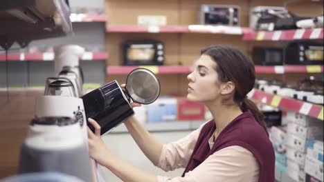 Young-female-costumer-choosing-new-electric-kettle-in-appliance-store-comparing-different-models-in-row-on-showcase.-Looking-for-kitchenware-in-department-of-household-electronics