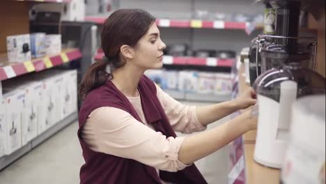 Young-female-costumer-examining-plastic-containers-for-electrical-juicer-looking-for-perfect-quality-kitchenware-in-appliance-store.-Household-equipment