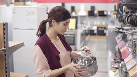 Una-Clienta-Joven-Eligiendo-Una-Olla-Entre-Una-Variedad-De-Utensilios-De-Cocina-En-Una-Tienda-De-Electrodomésticos.-Examinando-Cuidadosamente-Las-Bandejas-De-Acero-En-Una-Fila-De-Vitrinas.