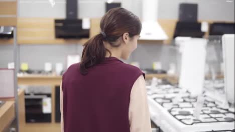 Young-girl-in-her-20's-searching-for-perfect-cooktop-in-hardware-store.-She-checks-the-price-and-opens-the-oven-of-a-white-cookstove