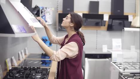 Attractive-brunette-girl-in-her-20's-looking-for-stove-hood-in-hardware-store-comes-up-to-white-set-and-checks-out-the-price