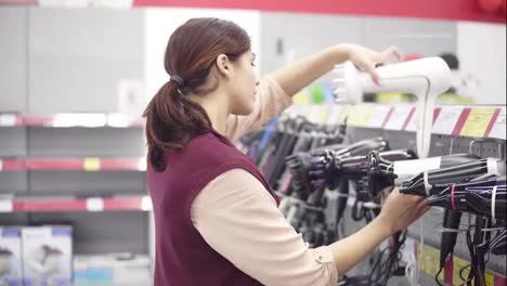 Young-beautiful-brunette-girl-picks-up-white-hairdryer-from-display-shelf-checks-it-out-and-puts-it-back-in-appliance-store.-Choosing-blow-dryer.-Household-equipment