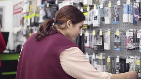 Joven-Y-Atractiva-Chica-Morena-Tratando-De-Escoger-Auriculares-Leyendo-Etiquetas-En-Envases-En-Exhibición-En-Una-Tienda-De-Electrónica