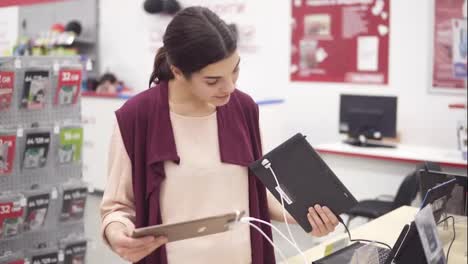 Young-beautiful-brunette-girl-smiling-while-choosing-between-two-tablets-grey-and-gold-attached-to-a-display-shelf-in-electronics-store