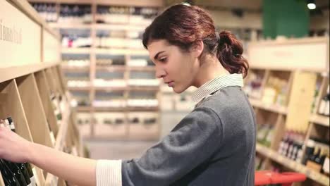 Young-beautiful-brunette-caucasian-girl-compares-two-different-bottles-of-wine.-Choosing-white-wine-at-grocery-store