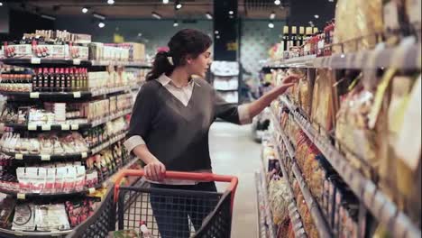 Young-brunette-girl-picks-out-two-packs-of-macaroni-or-pasta-at-local-grocery-store.-Shopping-push-cart-full-of-products,-no-lables-or-brands-visible.