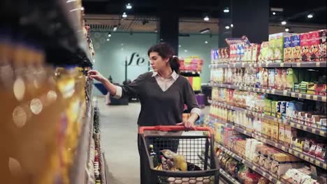 Beautiful-brunette-girl-walking-through-the-aisle-in-a-grocery-store-trying-to-choose-what-sause-to-buy.-Picking-glass-bottles