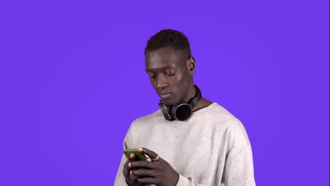 Un-Joven-Afroamericano-Que-Lleva-Auriculares-En-El-Cuello-Está-Escribiendo-En-Su-Teléfono-Inteligente-Mientras-Se-Encuentra-Aislado-Sobre-Un-Fondo-De-Pared-Azul.