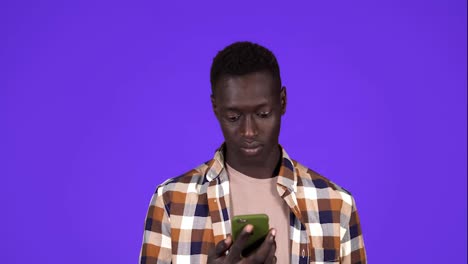 Young-black-man-holding-a-phone-crossing-fingers-for-having-luck,-looking-on-mobile-screen-and-burst-in-smiling,-winner