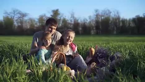 Happy-family,-father-and-daughters-are-sitting-close-together-and-enjoy-the-time-at-the-picnic-on-the-green-beautiful-meadow-on