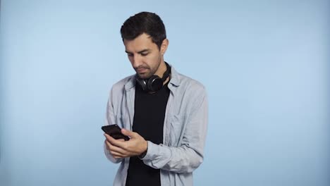 Young-confident-man-holding-phone-and-scroll-screen-or-typing-message-isolated-on-blue-background.-Wearing-headphones-on-neck