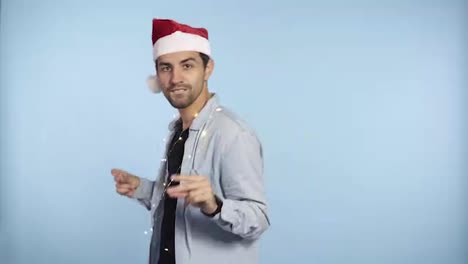 Positive-handsome-man-in-a-Christmas-redhat-and-garland-on-neck-dances-and-smiles-at-the-camera---blue-wall-background-studio