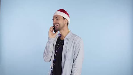 Young-happy-emotional-man-wearing-christmas-santa-hat-and-garland-on-neck-standing-and-talking-by-mobile-phone.-Man-in-casual