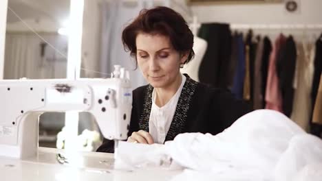 Portrait-of-and-adult-woman-seamstress-works-at-a-sewing-machine,-working-on-a-white-handmade-dress.-Fashion-clothes-on-the