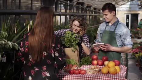 Cheerful-saleswoman-with-her-husband-is-putting-fresh-parsley-in-paper-bag-to-customer-in-greenhouse-market-talking-smiling