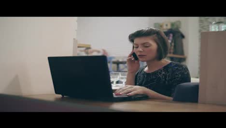 Young-business-woman-sitting-by-the-window-with-a-laptop-and-talking-on-the-phone