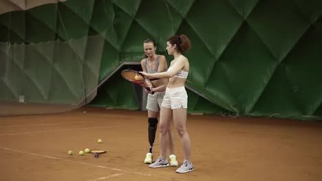 Detrás-De-Ella,-Una-Bella-Entrenadora-Con-Una-Prótesis-En-La-Pierna-Muestra-A-Su-Pupilo-Los-Matices-Del-Golpe-De-La-Pelota-En-El-Tenis.-Chicas-Deportivas