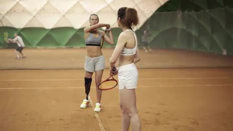 A-beautiful,-athletic-invalid-girl-teaches-the-beginner-of-the-correct-trajectory-of-a-racket-hit-in-tennis.-Other-players-on-the-background.-Slow-motion