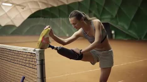 A-slender-disabled-girl-stretches-her-injured-leg-on-the-tennis-net-before-the-game.-Prosthesis.-Indoors