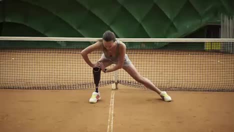 A-beautiful-girl-stretches-her-legs-muscles-on-and-indoor-tennis-court.-Disabled.-Tennis-net-behind.-Front-view