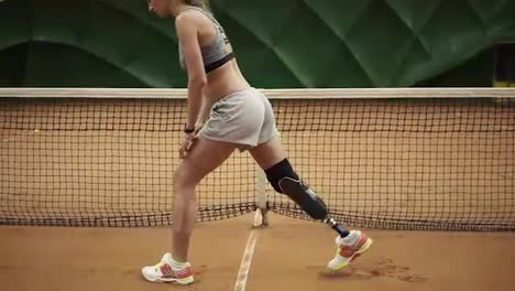 A-woman-with-a-prosthesis-stretches-her-legs,-making-lunges-against-the-background-of-the-tennis-court.-Sportswear.-Slow-motion