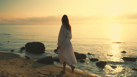 Lonely-Woman-In-White-Dress-Walking-Barefooted-On-Rocky-Seaside-During-Dusk