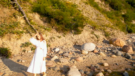 A-young-woman-in-a-white-dress-walks-along-the-edge-of-a-sandy-beach-with-a-few-large-rocks-during-sunrise---in-the-background,-a-cliff-descending-to-the-sea