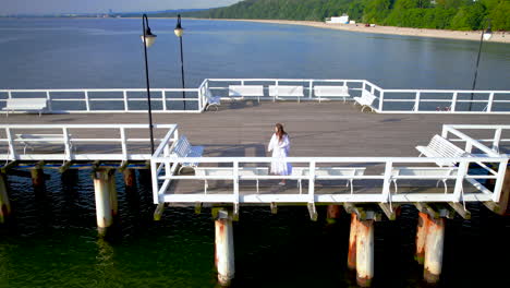 Young-Woman-in-White-Dress-Observing-Scenic-Seascape-View-Walking-on-Orlowo-Pier,-Gdynia,-Poland,-Aerial-shot
