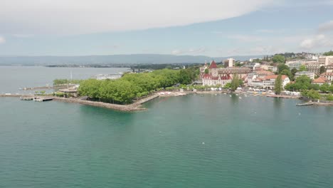 Drone-flying-away-from-the-beautiful-city-of-Lausanne-at-the-shoreline-of-Lake-Geneva,-Switzerland