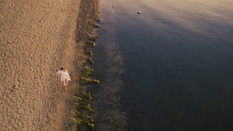 Woman-In-White-Summer-Dress-Walking-By-the-Sea-Along-Sandy-Beach-Coastline-at-Sunrise---Aerial-Tracking-Top-Down