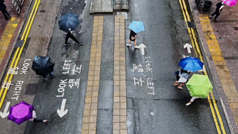 Vista-De-Arriba-Hacia-Abajo-De-Personas-Con-Sombrillas-Cruzando-La-Calle-En-Hong-Kong-Lluvioso