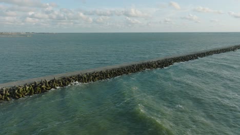Aerial-establishing-view-of-Port-of-Liepaja-concrete-pier,-Baltic-sea-coastline-,-sunny-day-summer-evening,-golden-hour-light,-big-waves-splashing,-wide-drone-dolly-shot-moving-right