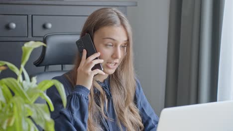 Young-woman-looking-surprised-while-on-phone-while-working-with-laptop-at-office