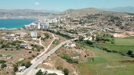 Suburbs-of-Saranda-city,-cars-driving-on-the-road-between-mountains-and-the-sea