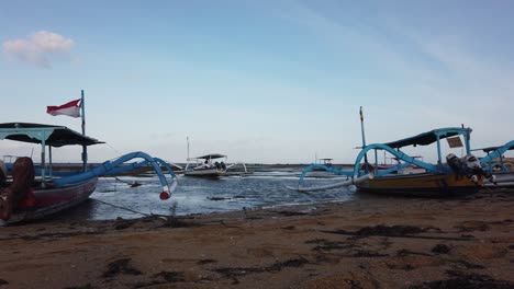 Traditionelle-Hölzerne-Segelboote,-Die-Am-Strand-Von-Sanur-Auf-Bali-Angedockt-Sind.-Die-Indonesische-Flagge-Weht-Im-Wind,-Sommer-In-Südostasien