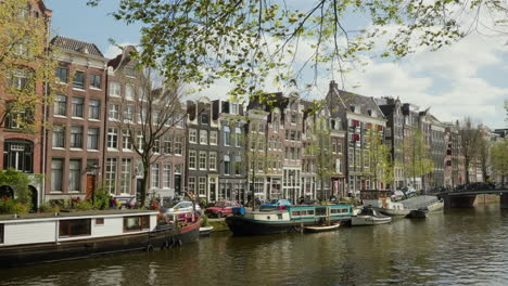 Eine-Ruhige-Kanalszene-In-Amsterdam-Im-Frühling,-Gespickt-Mit-Hausbooten-Und-Klassischen-Gebäuden-Entlang-Der-Uferpromenade