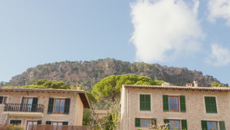 Houses-in-Valldemossa,-Mallorca,-set-against-a-backdrop-of-mountains,-blue-sky,-and-clouds,-portraying-a-beautiful-Spanish-town-scene
