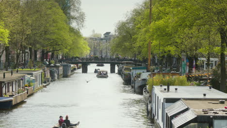 A-serene-Amsterdam-canal-in-Spring,-with-boats-leisurely-cruising-along-the-water-under-the-good-weather