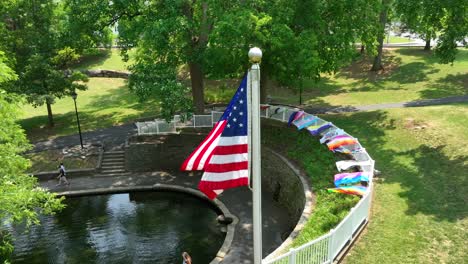 Panoramic-aerial-view-of-a-community-park-with-a-walking-path-around-a-lake,-pride-flags-attached-to-the-surrounding-fence-and-the-American-flag-blowing-gently-in-the-breeze