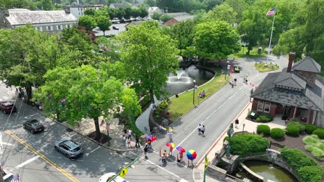 Aerial-view-of-a-community-preparing-for-a-pride-festival,-with-a-fountain-in-the-lake-in-the-park,-people-holding-pride-umbrellas,-an-American-flag-blowing-gently-in-the-breeze-and-a-pavilion