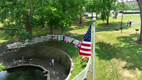 Aerial-view-of-a-community-park-with-people-out-walking-around-a-lake,-pride-flags-attached-to-the-surrounding-fence-and-the-American-flag-blowing-gently-in-the-wind