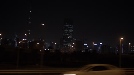 Mit-Dem-Auto-Fahren-Und-Nachts-In-Einem-Auto-Auf-Der-Autobahn-An-Der-Skyline-Von-Dubai-Vorbeifahren,-Mit-Entfernten,-Beleuchteten-Gebäuden-Und-Wolkenkratzern,-Darunter-Das-Burj-Khalifa-In-Den-Vereinigten-Arabischen-Emiraten