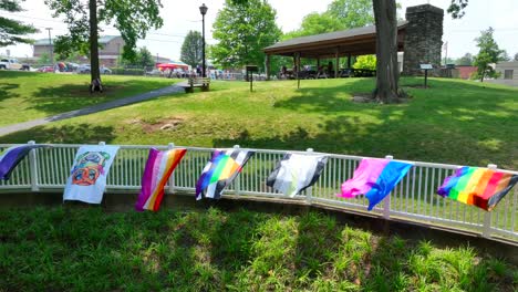 Drone-footage-of-a-pride-festival-being-held-at-a-community-park-with-pride-flags-attached-to-a-fence,-a-pavilion-and-people-congregating-on-a-beautiful-day
