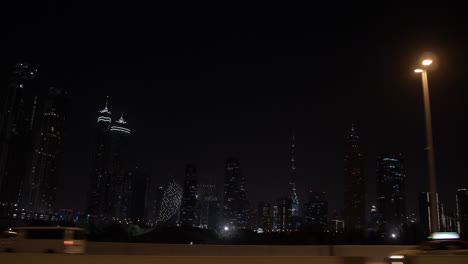 Passing-by-the-Dubai-skyline-at-night-in-a-car-on-the-highway-with-distant,-lit-up-buildings-and-skyscrapers-including-the-Burj-Khalifa-in-the-United-Arab-Emirates