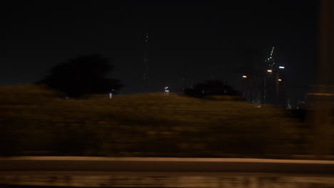 Fahren-Sie-Nachts-In-Einem-Auto-Auf-Der-Autobahn-An-Der-Skyline-Von-Dubai-Vorbei,-Mit-Entfernten,-Beleuchteten-Gebäuden-Und-Wolkenkratzern,-Darunter-Das-Burj-Khalifa-In-Den-Vereinigten-Arabischen-Emiraten