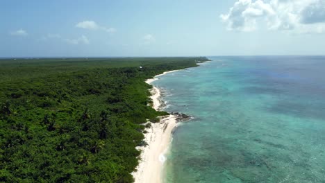 Drone-footage-flying-backwards-above-a-turquoise-ocean-and-coral-reef-in-the-Caribbean-as-native-forest-stretches-along-a-beach-into-the-horizon-and-clouds-cast-shadows-over-the-water