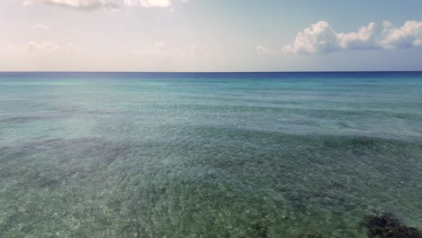 Drone-footage-flying-out-over-a-turquoise,-tropical-ocean-as-waves-crash-over-a-coral-reef-and-onto-a-beach-in-the-Caribbean-