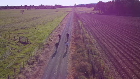 Flying-over-a-group-of-cyclists-riding-along-a-straight-gravel-road-in-farmland,-New-Zealand