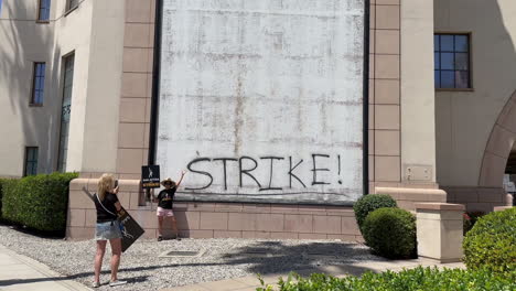SAG-AFTRA-Striker-with-Picket-Sign-posing-for-photo-in-front-of-STRIKE-Graffiti-by-Warner-Brothers-Studios-on-Olive-Ave-Burbank-CA-at-around-12PM